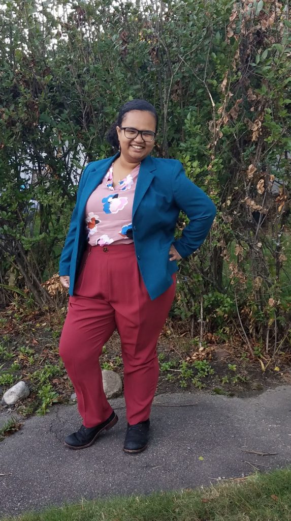 Talia Franks, a light skinned Black person with brown eyes, blue glasses, and black hair slicked back into a curly pony tail grins at the camera with one hand at their side and the other at their hip while standing in front of a large bush. They are wearing a pink shirt with blue, red, and pink flowers and a blue blazer with waist high red pants and black boots.