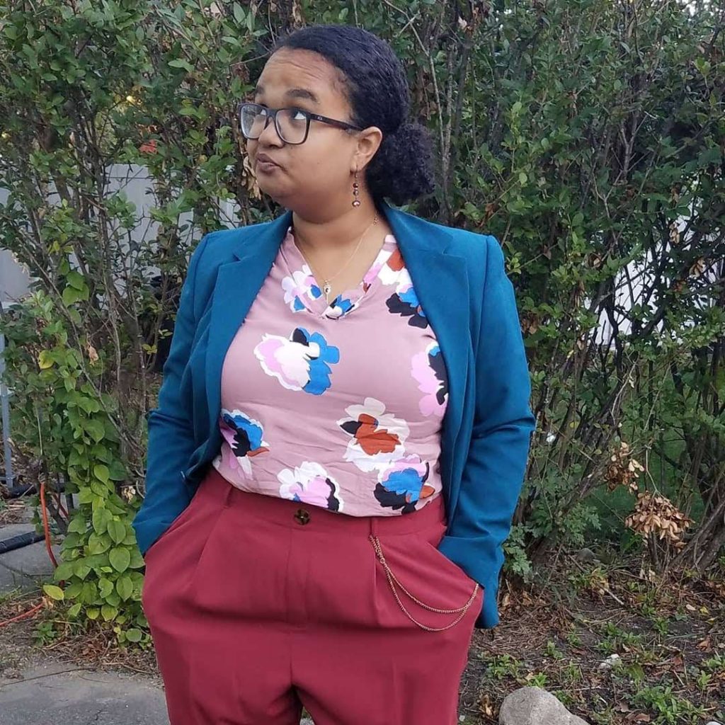 Talia Franks, a light skinned Black person with brown eyes, blue glasses, and black hair slicked back into a curly pony tail smirks into the distance with their hands in their pockets while standing in front of a large bush. They are wearing a pink shirt with blue, red, and pink flowers and a blue blazer with waist high red pants that have a pocket watch chain.
