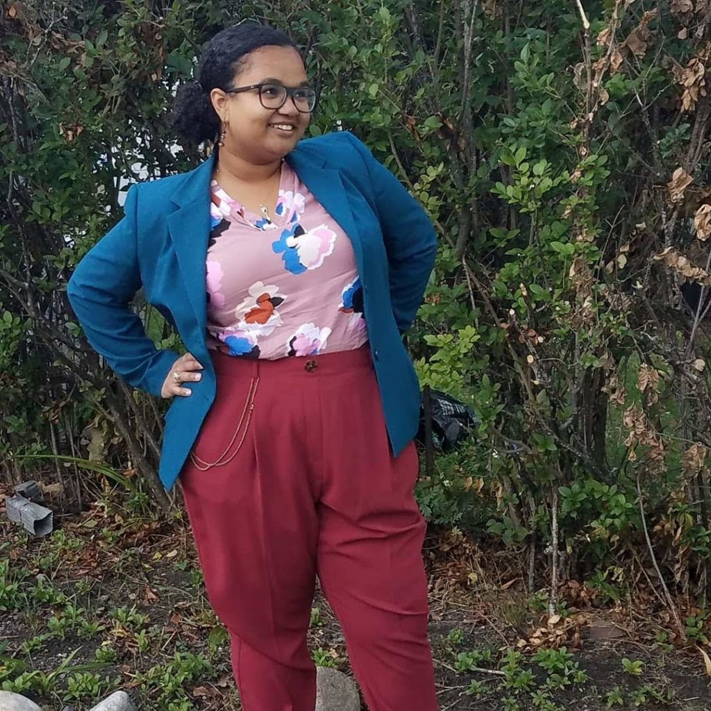 Talia Franks, a light skinned Black person with brown eyes, blue glasses, and black hair slicked back into a curly pony tail smiles into the distance with their hands on their hips while standing in front of a large bush. They are wearing a pink shirt with blue, red, and pink flowers and a blue blazer with waist high red pants that have a pocket watch chain.