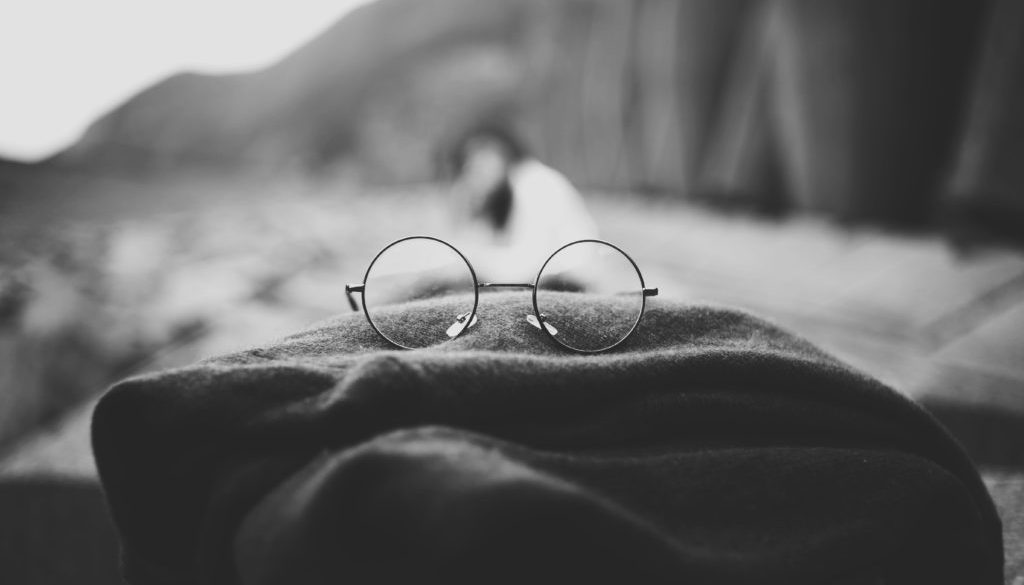 Grayscale photo of round glasses placed on top of a folded scarf with a figure and some cliffs blurred in the background, perhaps on a beach.