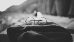Grayscale photo of round glasses placed on top of a folded scarf with a figure and some cliffs blurred in the background, perhaps on a beach.