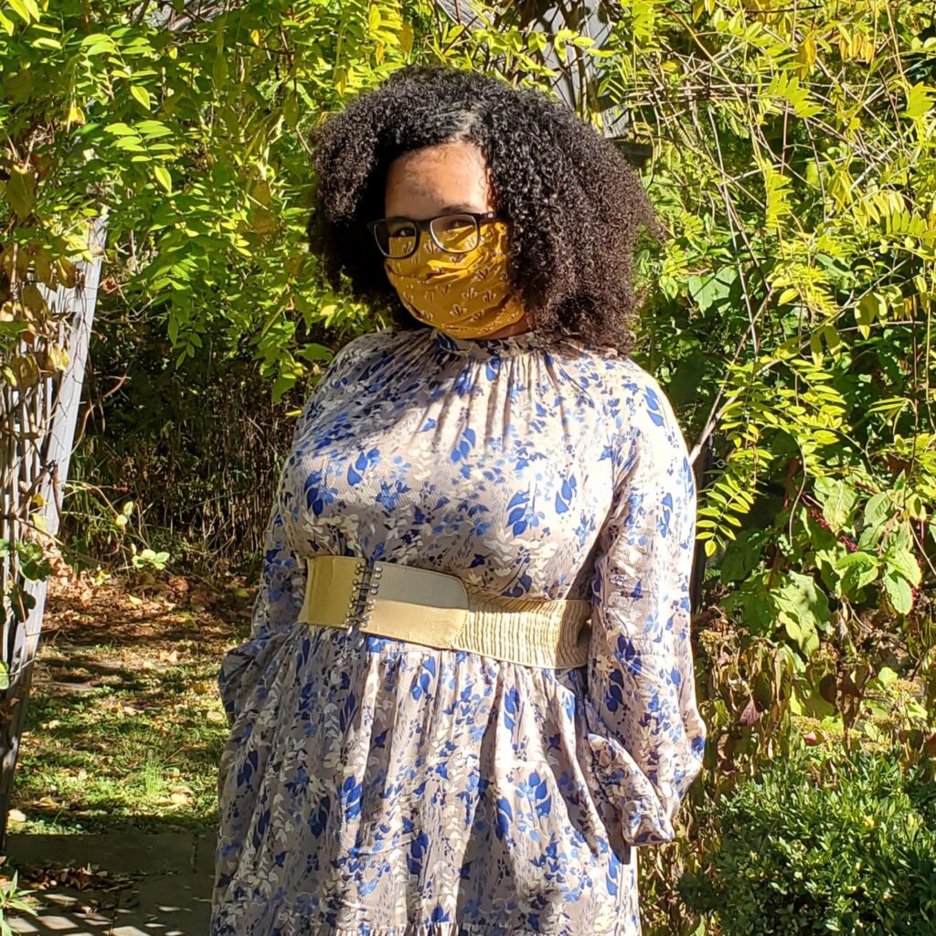 Talia Franks, a light skinned Black person with curly black hair, blue glasses, and a yellow mask looks into the camera. They are wearing a blue and white dress with flowers and a gold belt, and standing in a garden in front of a blue archway covered in ivy