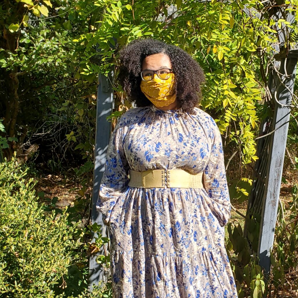 Talia Franks, a light skinned Black person with curly black hair, blue glasses, and a yellow mask looks into the distance. They are wearing a blue and white dress with flowers and a gold belt, and standing in a garden in front of a blue archway covered in ivy