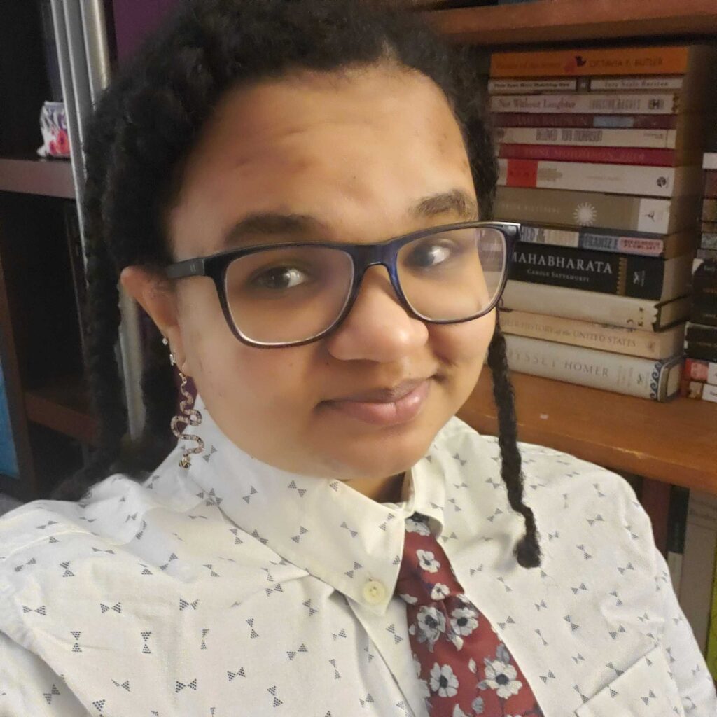 Talia Franks, a lightskinned Black person with brown eyes, black hair in three large braids, and blue glasses smiles in a selfie. They are wearing a white button down shirt with blue bowties and a red necktie with white and blue flowers and are sitting in front of a bookshelf stacked with nonfiction and fiction books