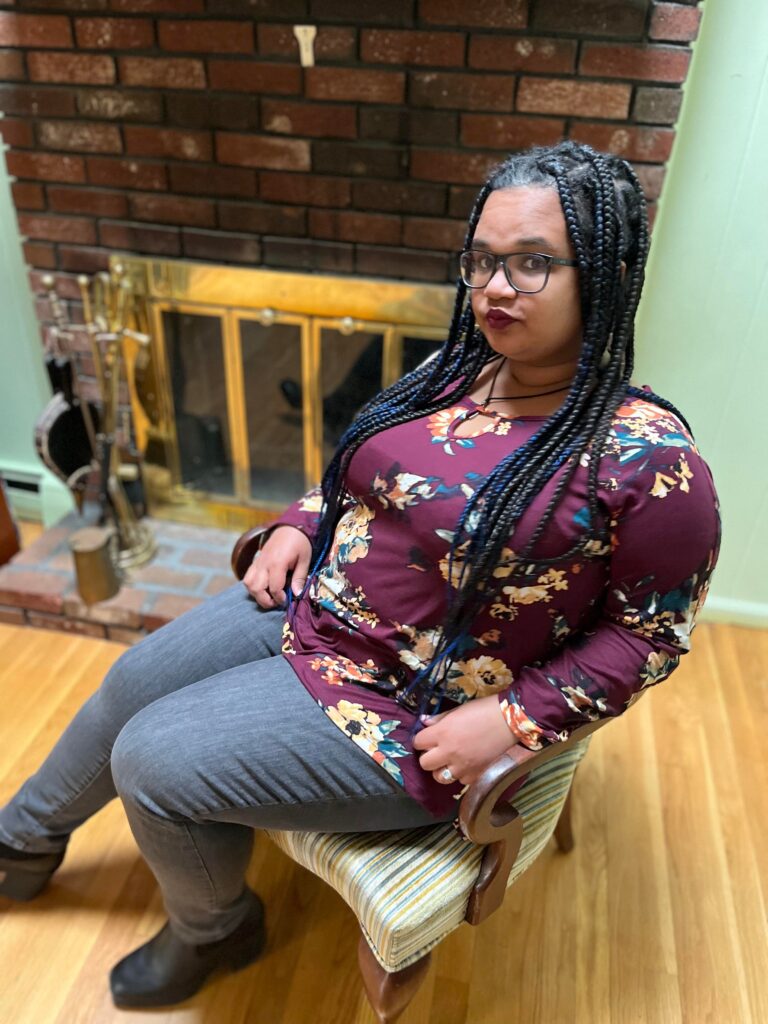 Talia Franks, a lightskinned Black person with brown eyes, long knotless braids and large blue glasses sits in front of a fireplace.