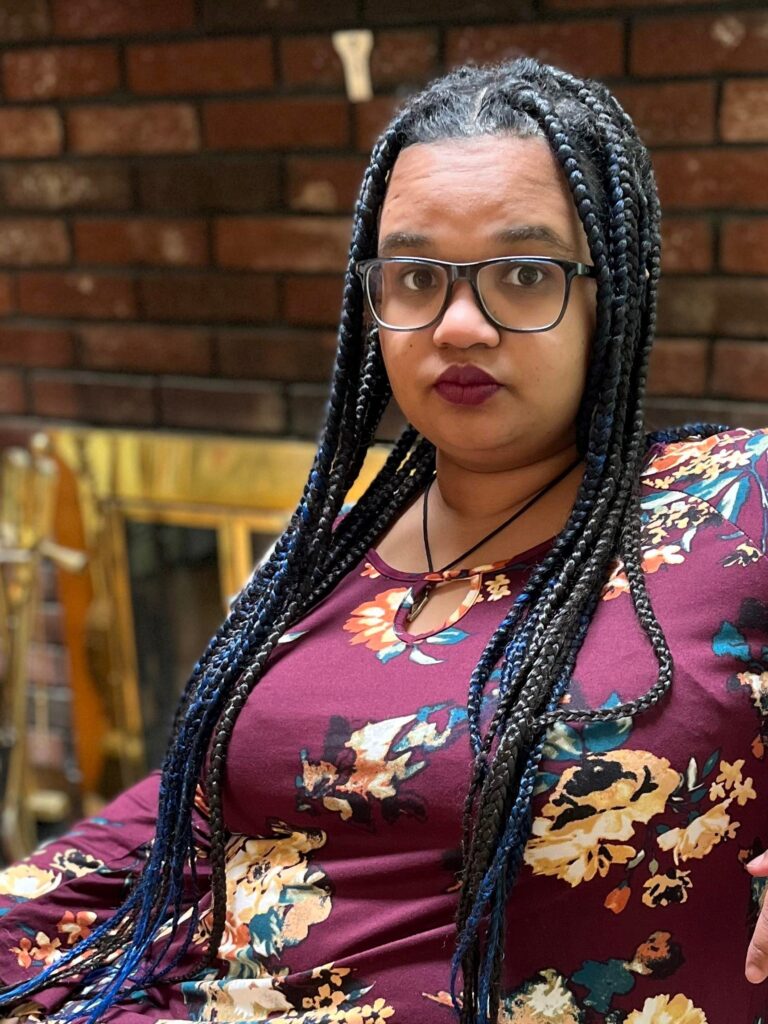 Talia Franks, a lightskinned Black person with brown eyes, long knotless braids and large blue glasses sits in front of a fireplace.