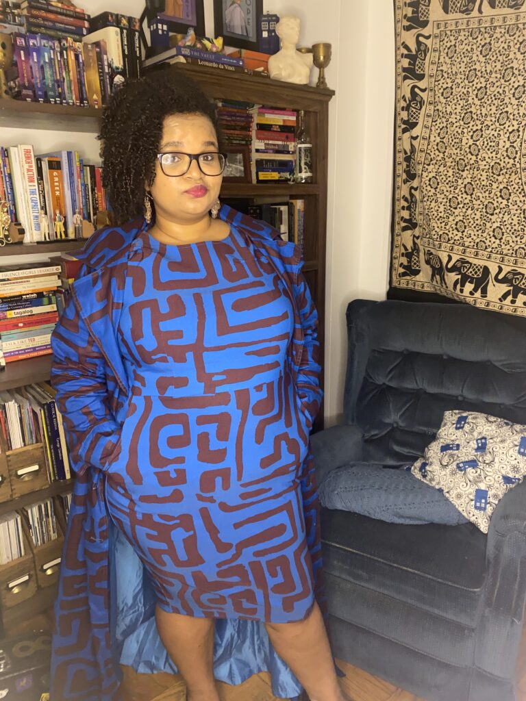 Talia in a blue and brown patterned dress with matching trenchcoat standing in front of a set of bookshelves and blue armchair