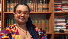 Selfie of Talia in front of three bookcases. They are wearing a kente cloth shirt and have their hair in knotless braids