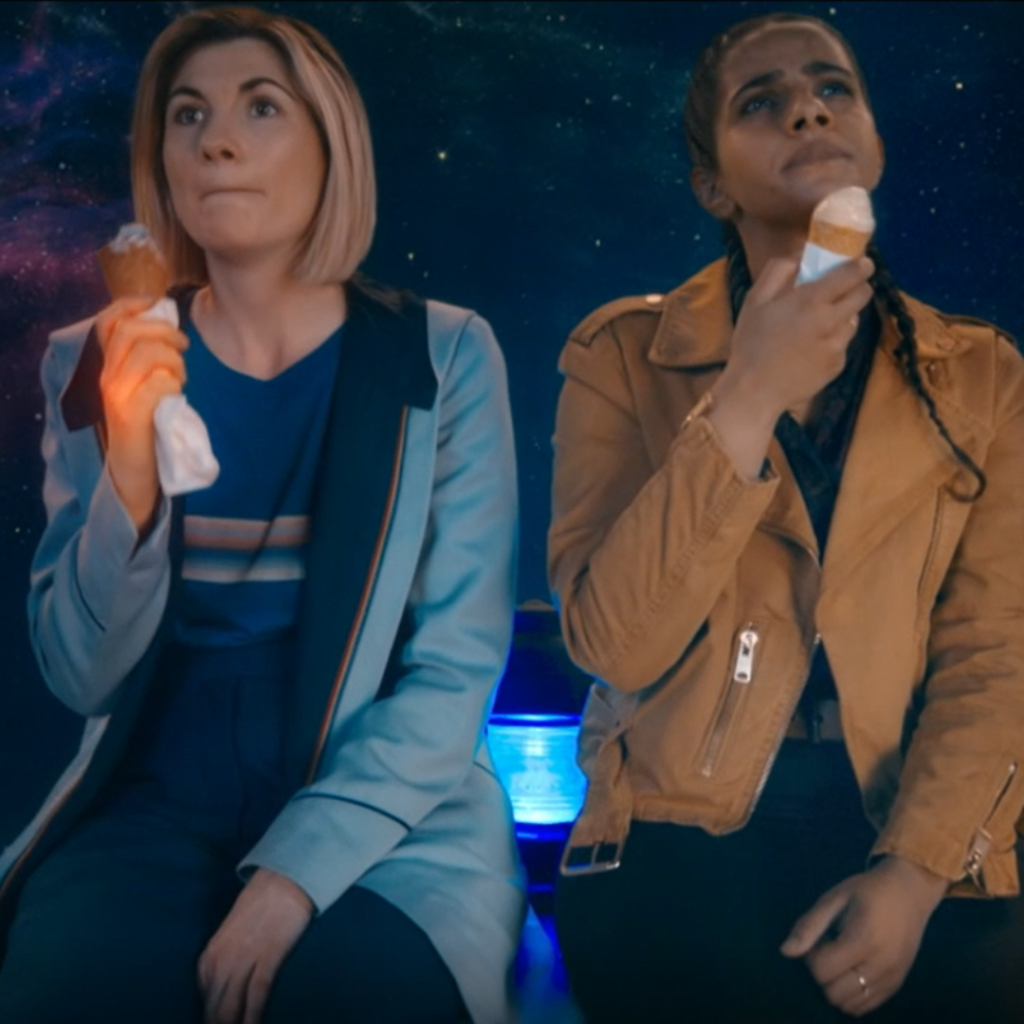 The Thirteenth Doctor (Jodie Whittaker) and Yaz (Mandip Gill) sitting on top of the Tardis with ice cream cones as it orbits in space. The Doctor's hand is glowing with regenerative energy.