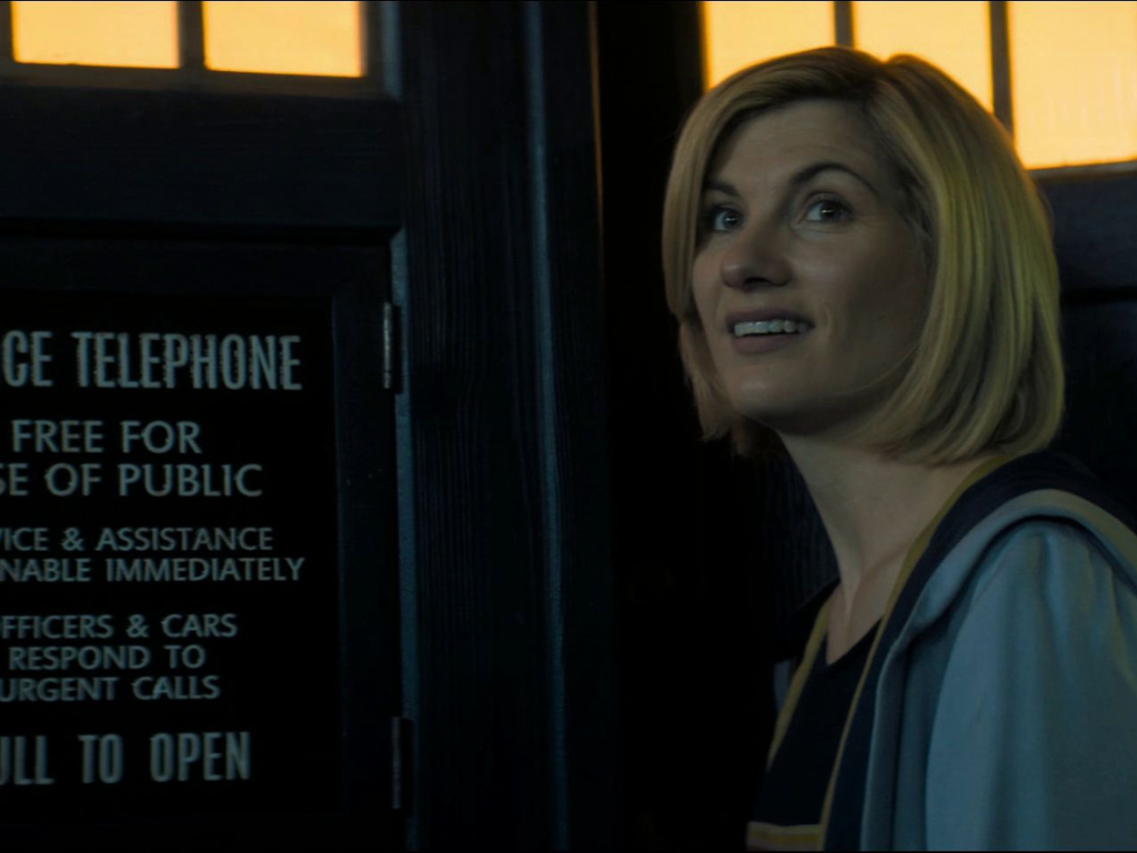 The Thirteenth Doctor (Jodie Whittaker) smiling while standing in the doorway of the Tardis